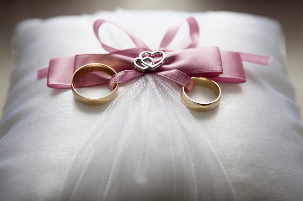 Close-up of elegant wedding rings on a silk pillow with a pink ribbon and a heart decoration, symbolizing love.